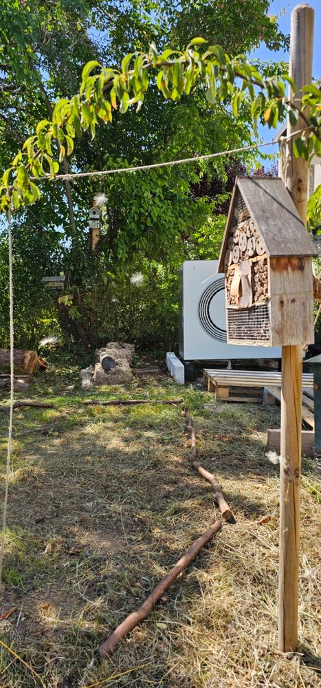 Eine Spinnenwebe zwischen rechts ein Insektenhotel, oben und links von einer Schnur gerahmt, hat ein paar weiße Pollenzeug. Im Hintergrund die Wärmepumpe und eine Bienen Wand mit Totholz.