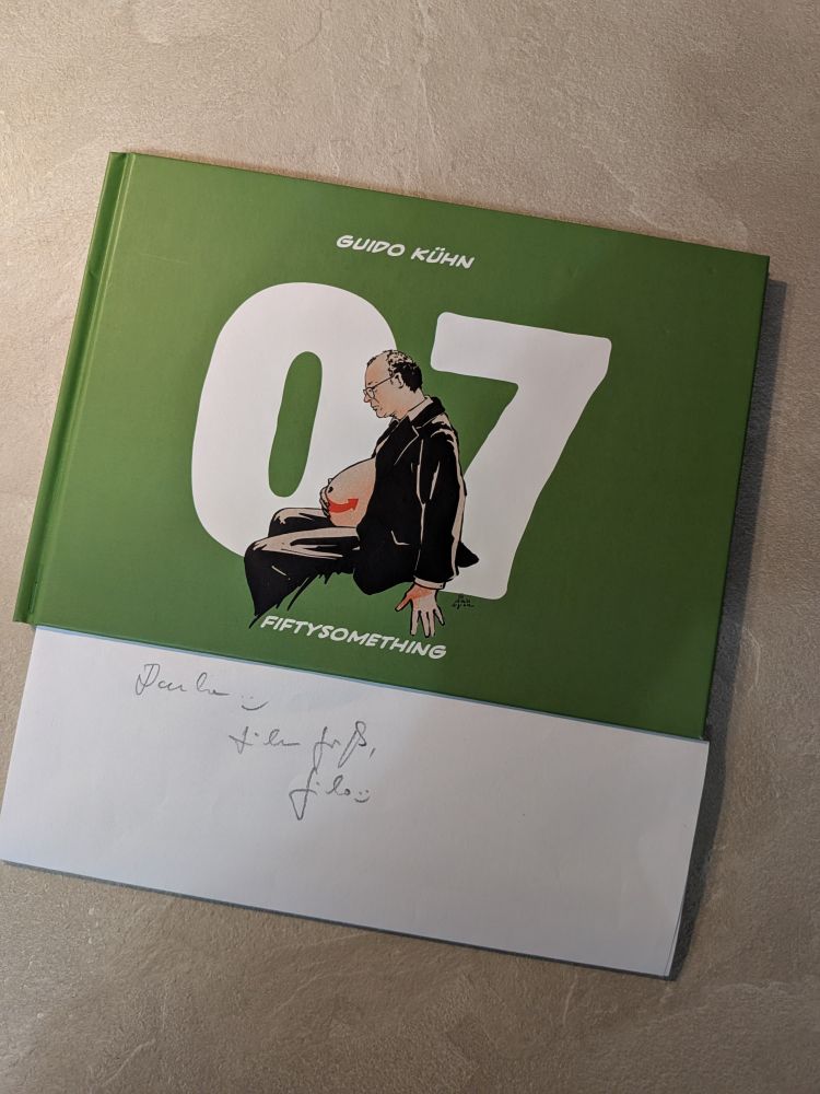 Ein Foto zeigt das Buch „07 – Fiftysomething“ von Guido Kühn mit grünem Hardcover. Auf dem Cover sitzt ein Mann mit Glatze im Profil, der eine Babytrage mit der Zahl 07 vor dem Bauch trägt. Darunter liegt ein Blatt mit handschriftlicher Widmung: „Vielen Dank, Guido :)“.
