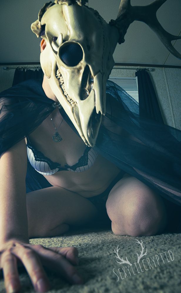 A deer skull-headed humanoid is crouched with one hand in the foreground and the rest of them in the background. They are wearing a black and white striped bra, black lace underwear, and a translucent cloak.