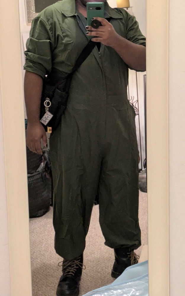 the same man with dark skin in front of a mirror, wearing a green jumpsuit, now worn normally, with the sleeves rolled up to the elbow.
he is also wearing black boots with yellow laces.