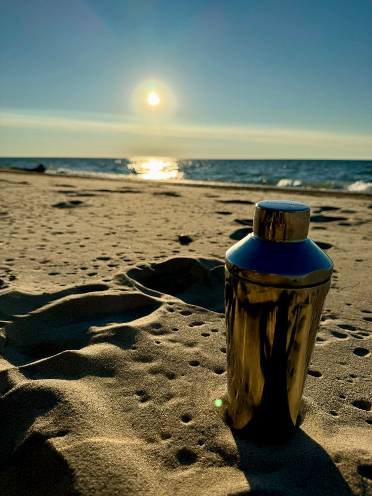 Martini shaker in the sand on the beach.  The sand is a warm golden hue and in the background the sun is going down over calm water.