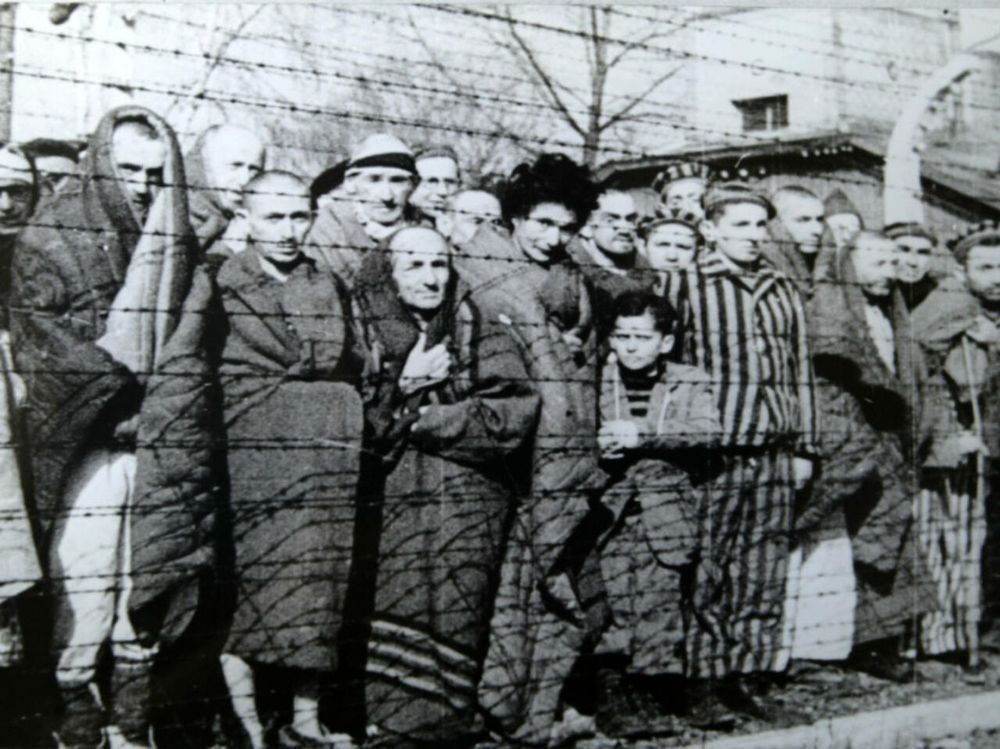 Photographie de déportés d’Auschwitz derrière des barbelés, certains enroulés dans une couverture, d’autres en pyjama rayé. De tous âges. 