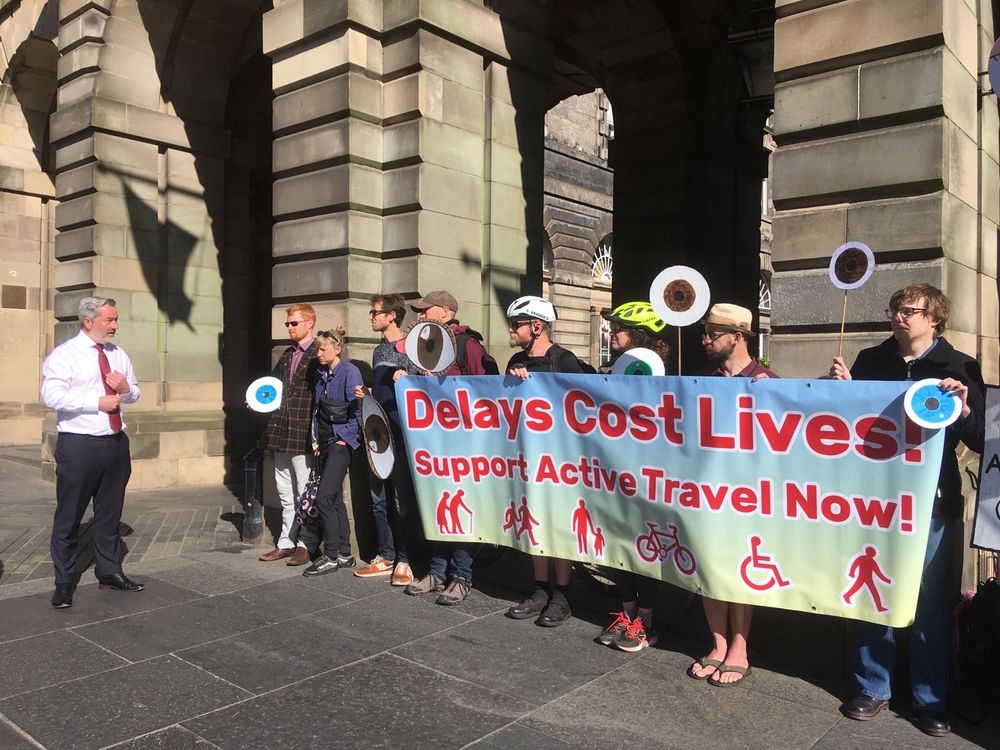 A group of people standing in front of the Edinburgh City Chambers. They are holding a big banner which says "Delays Cost Lives! Support active travel now!" and are holding lots of cardboard eyes. Some of them are talking to a man in a suit (Cllr Jenkinson).