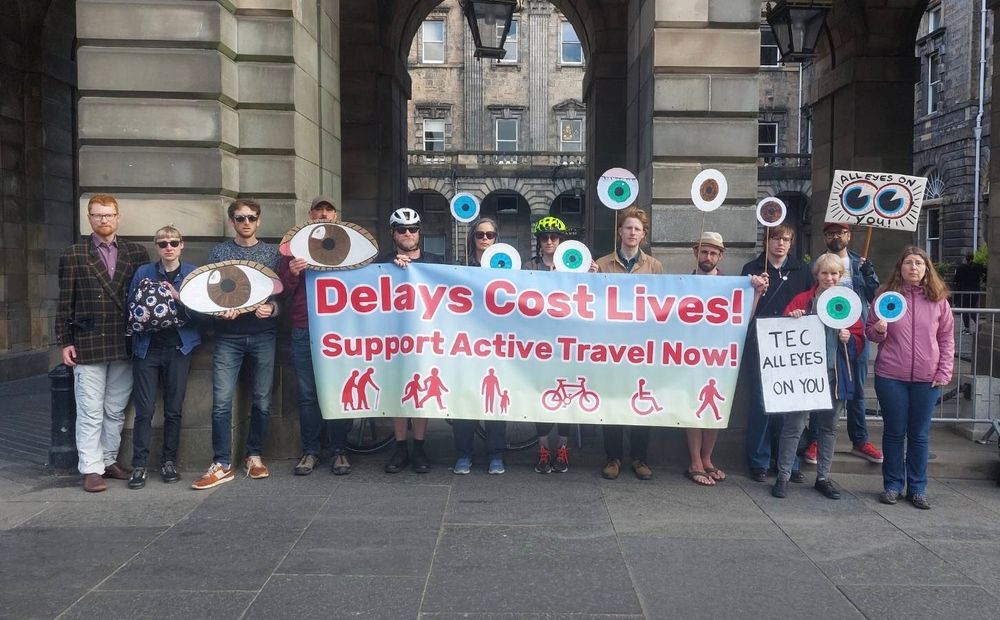 A group of people standing in front of the Edinburgh City Chambers. They are holding a big banner which says "Delays Cost Lives! Support active travel now!" and are holding lots of cardboard eyes.