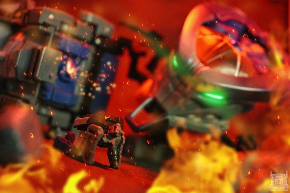 Photo edit of two Transformers figures. A titan master crawls on the ground after being ripped from the larger head vessel. Flames surround him as a large head-shaped insectoid tank looms in the background.