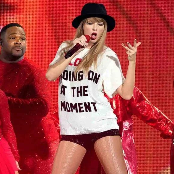 Taylor Swift at the eras tour wearing white “a lot going on at the moment” shirt and black hat and shorts holding a microphone 