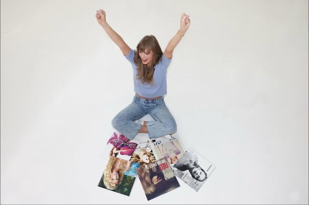 taylor swift happily sitting on the floor with her arms up in the air with her records in front of her 