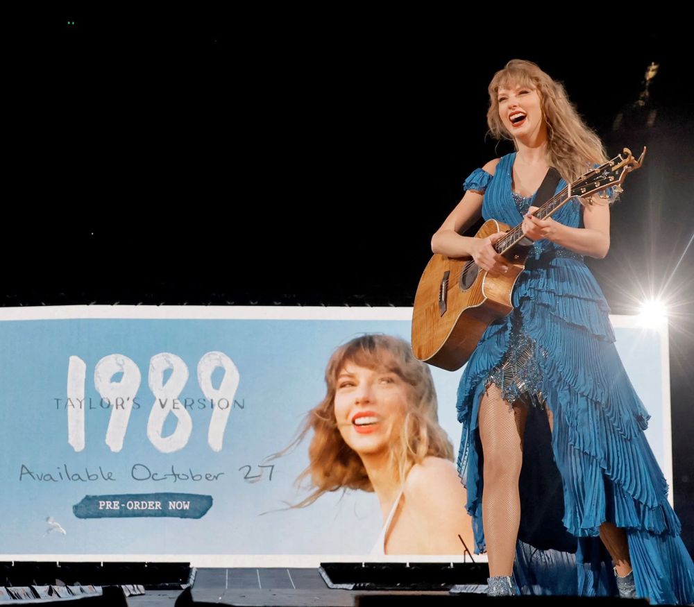 Taylor Swift wearing a blue dress holding a guitar during the surprise song set at the Eras Tour with the 1989 Taylor’s Version Announcement in the back 