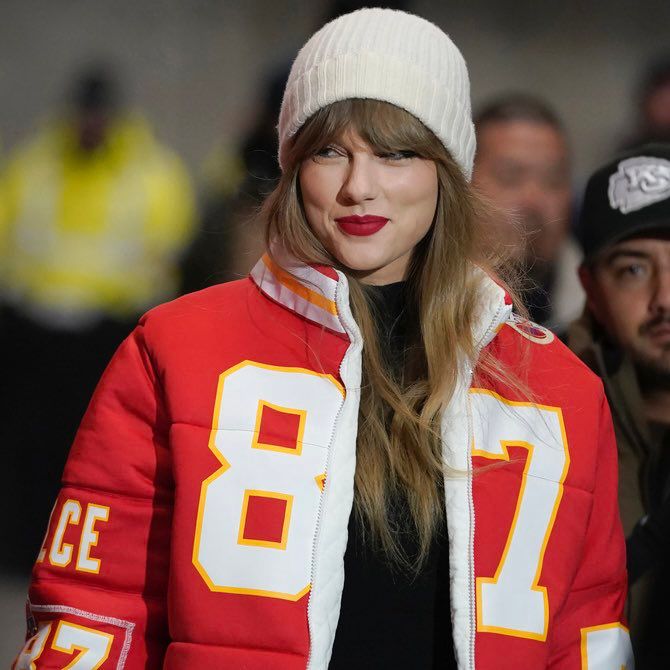 taylor swift wearing a black shirt and a puffy red, white, and yellow 87 jacket and a white hat at a chiefs game 