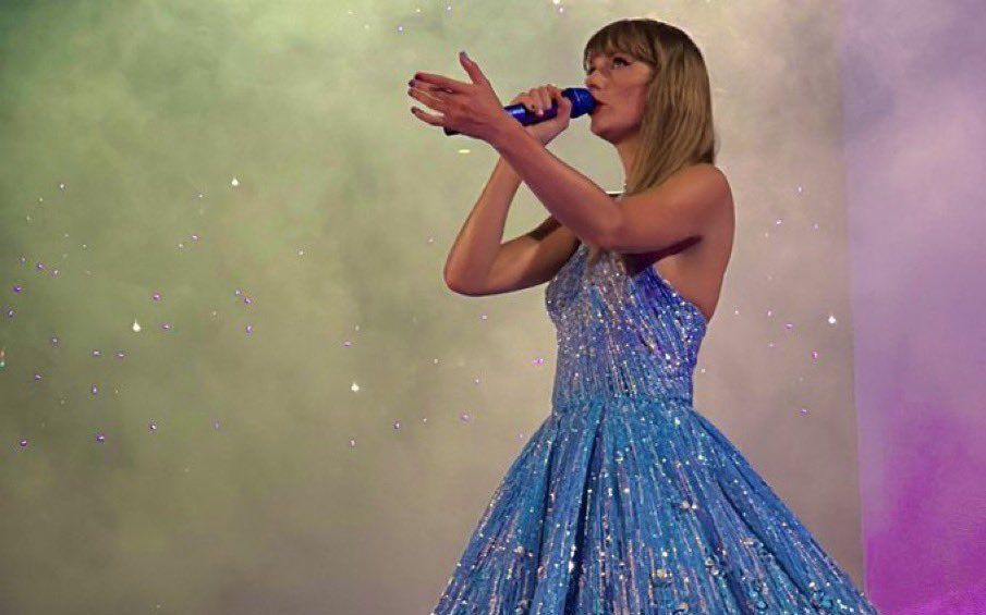 Taylor Swift in blue sparkly Speak Now dress holding a microphone at the Eras Tour 