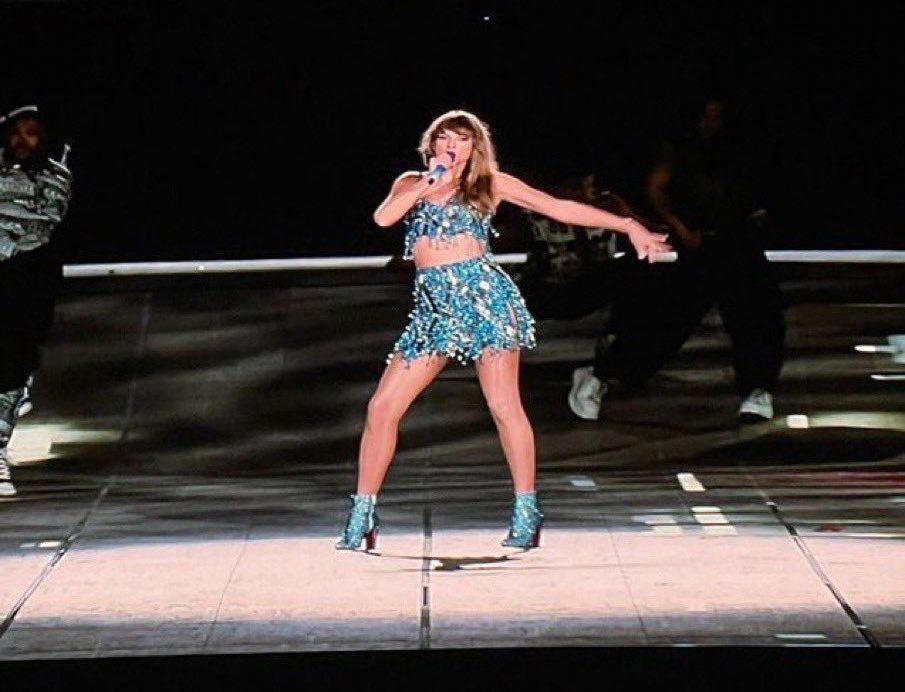 Taylor Swift in blue top, skirt, and boots holding a microphone during the 1989 set at the Eras Tour 