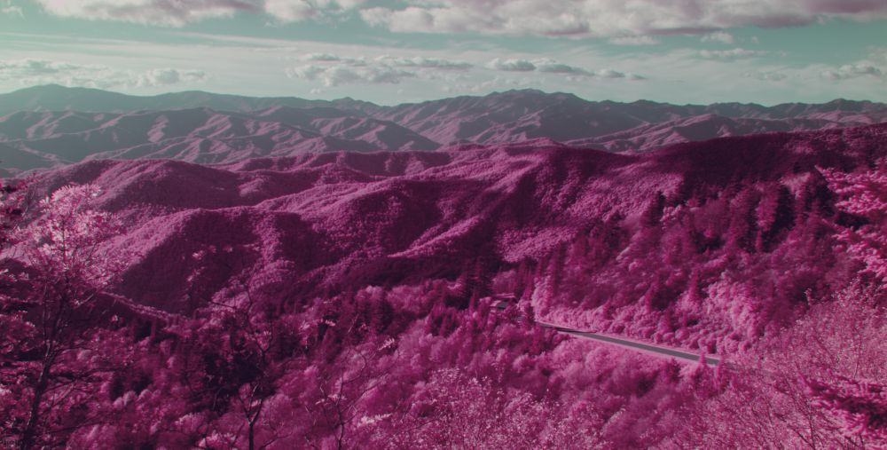 A bright pink infrared mountain landscape beneath a pale blue cloudy sky has a small portion of road winding around a hill on the right side.
