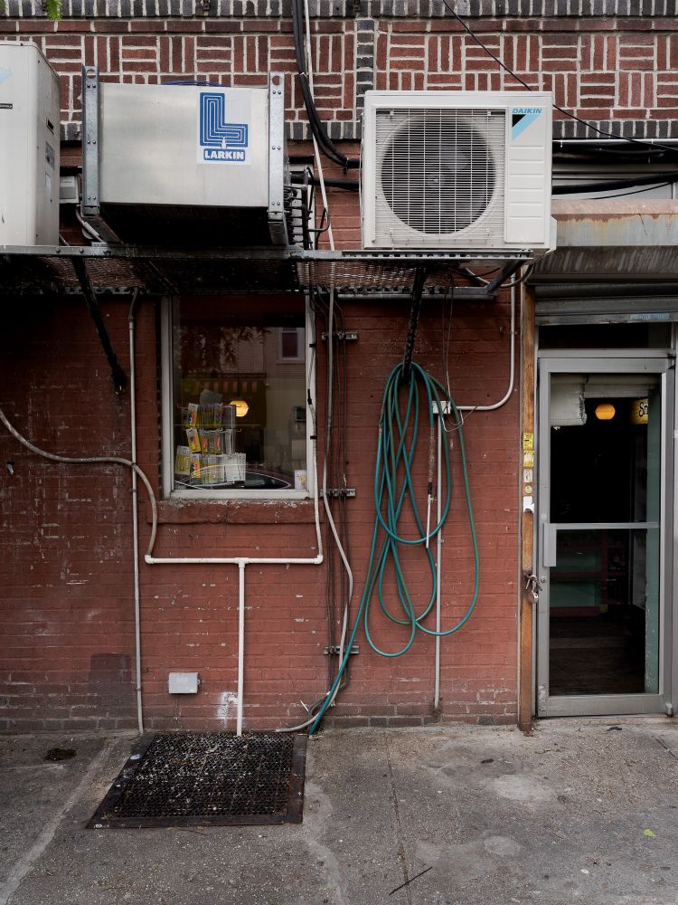 This is a color photograph of the back entrance to a retail shop in Brooklyn, New York. It features an interesting combination of hoses, pipes, ac units, windows and doors. 