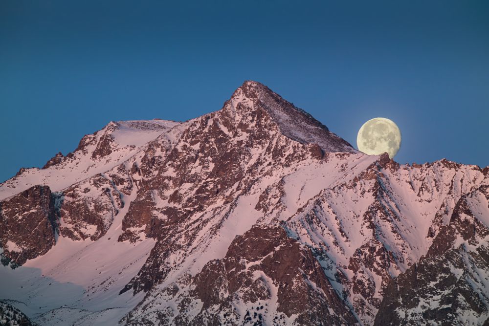 A snowy mountain peak at sunrise with the moon setting behind it.