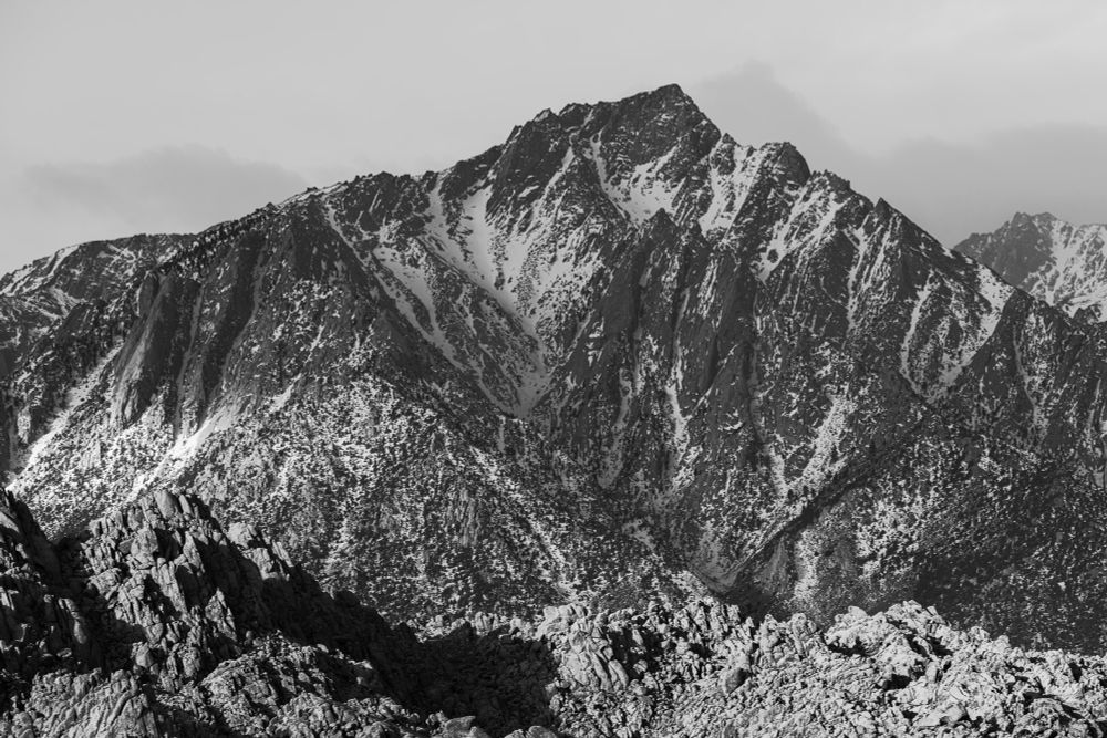 A dramatic black and white image of a rugged mountain range, featuring steep rocky peaks and patches of snow. The foreground includes jagged rock formations, while the background shows a towering peak against a cloudy sky.