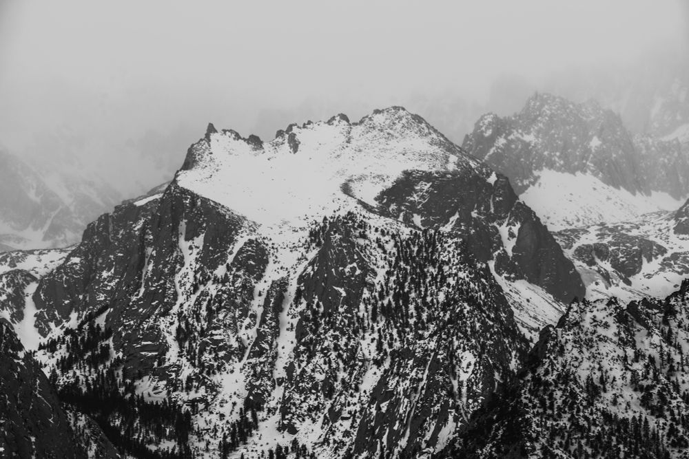 Black and white image of a rugged mountain landscape, featuring snow-covered peaks and rocky slopes, partially obscured by mist. Dense patches of trees are visible on the lower slopes.