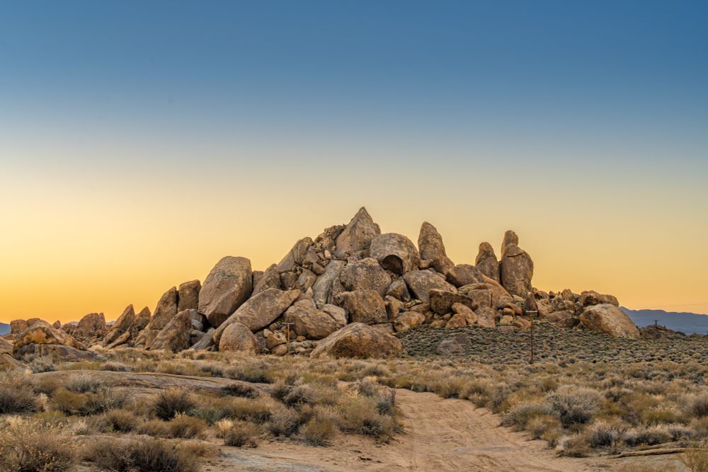 A rocky formation at sunrise, with a gradient sky transitioning from blue to orange. A dirt road leads towards boulders, surrounded by sparse vegetation.