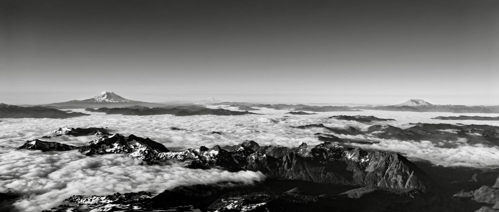 A black and white panoramic view of mountain ranges with snow-capped peaks above a sea of clouds. Several volcanic peaks are visible in the distance, fading into the horizon.