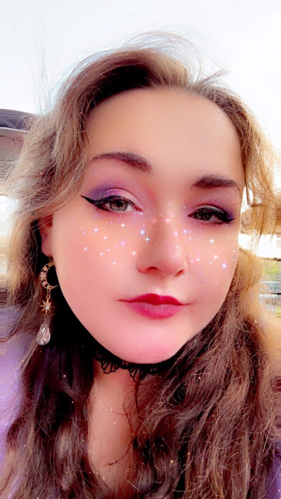 Selfie of me outside in the day with a moon dangle earring and Snapchat filter of stars across my nose and cheeks like freckles 