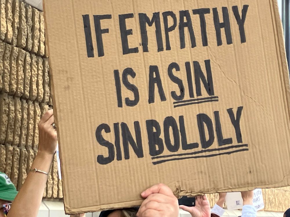 A hand holds up a handwritten sign that says: if empathy is a sin, sin boldly.