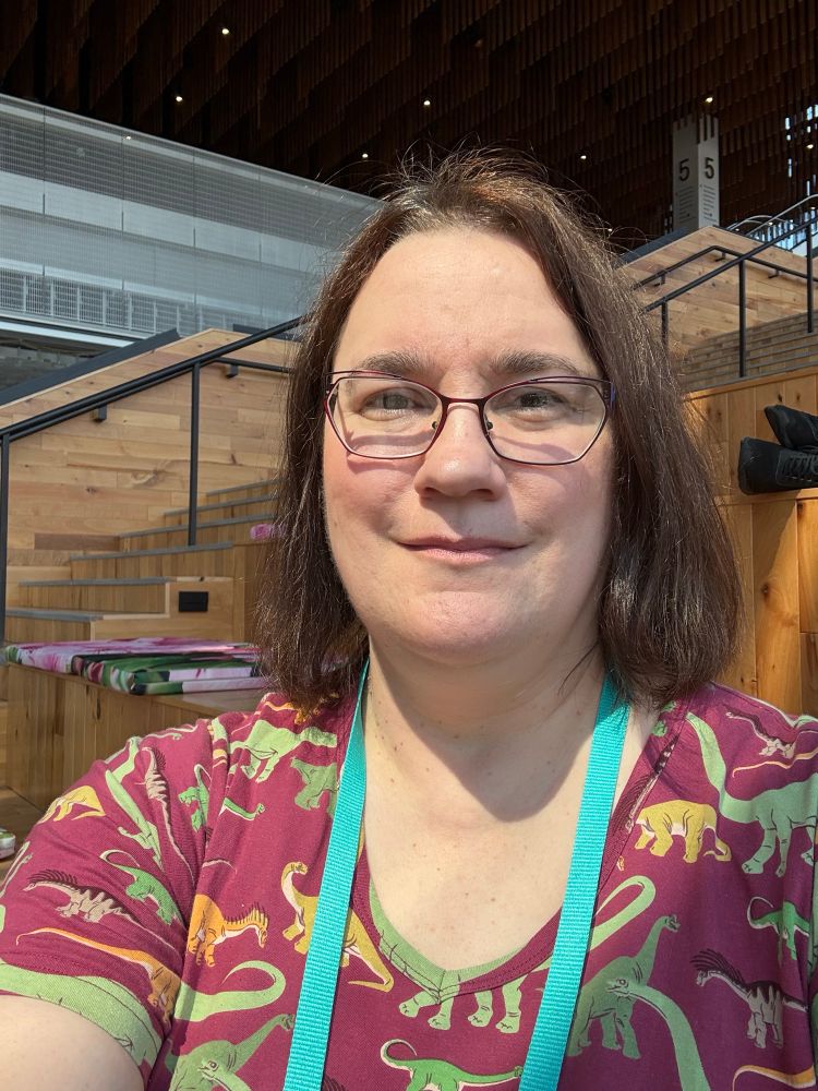 A middle-aged white woman with brown hair and glasses. Her dress is dinosaur print. 