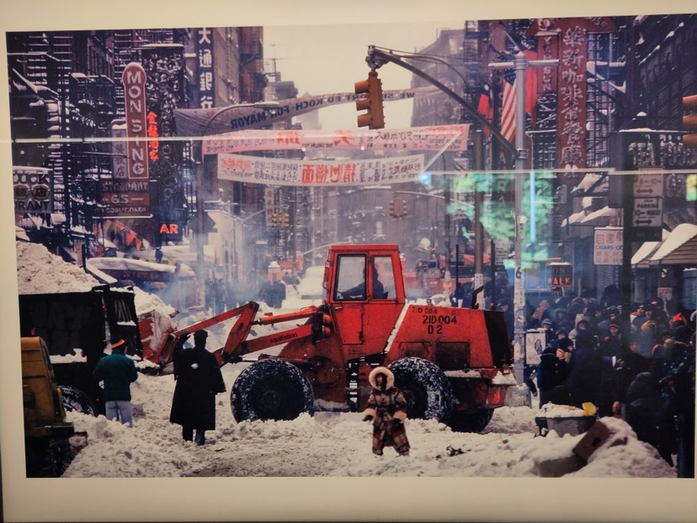 Man sieht eine Großstadt es ist eigentlich China Town in New York. Über der Straße hängen Transparente mit Kanjis. Es schneit sehr stark. In der Mitte steht ein roter Bagger, der Schnee schippen. Davor eine Person mit einem Mantel im Inuit-Stil: Fellkapuze, beige, braun und schwarz gemustert. Die Person steht knietief im Schnee