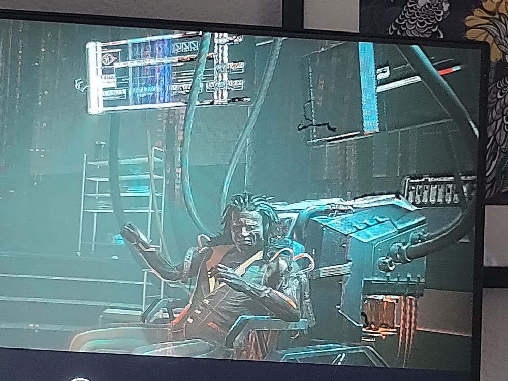 Man sieht eine Figur die verkabelt auf einem Stuhl sitzt. Der Mann hat einen Anzug und Dreadlocks. Das Bild ist in sehr kühlen Farben (blau, silber, grün schimmernd) Der Stuhl verbindet den Mann mit mehreren Monitoren