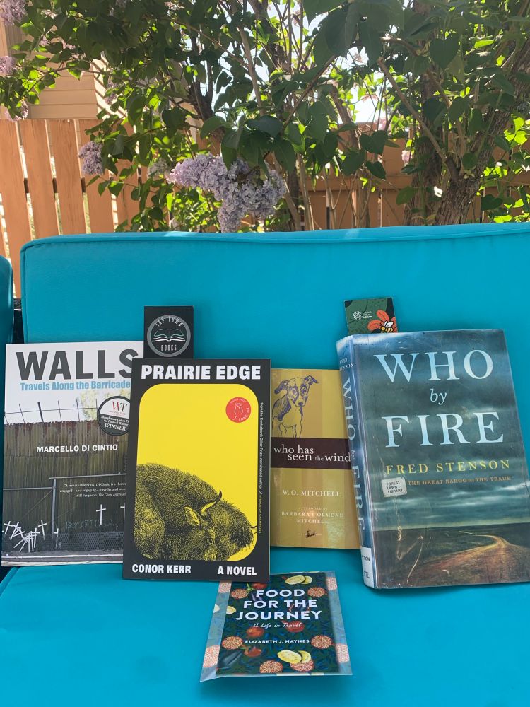 A selection of great books by Alberta authors with lilacs in the background - Walls by Marcello di Cintio, Prairie Edge by Conor Kerr, Who Has Seen the Wind by W.O. Mitchell, Who by Fire by Fred Stenson, Food for the Journey by Elizabeth J. Haynes.