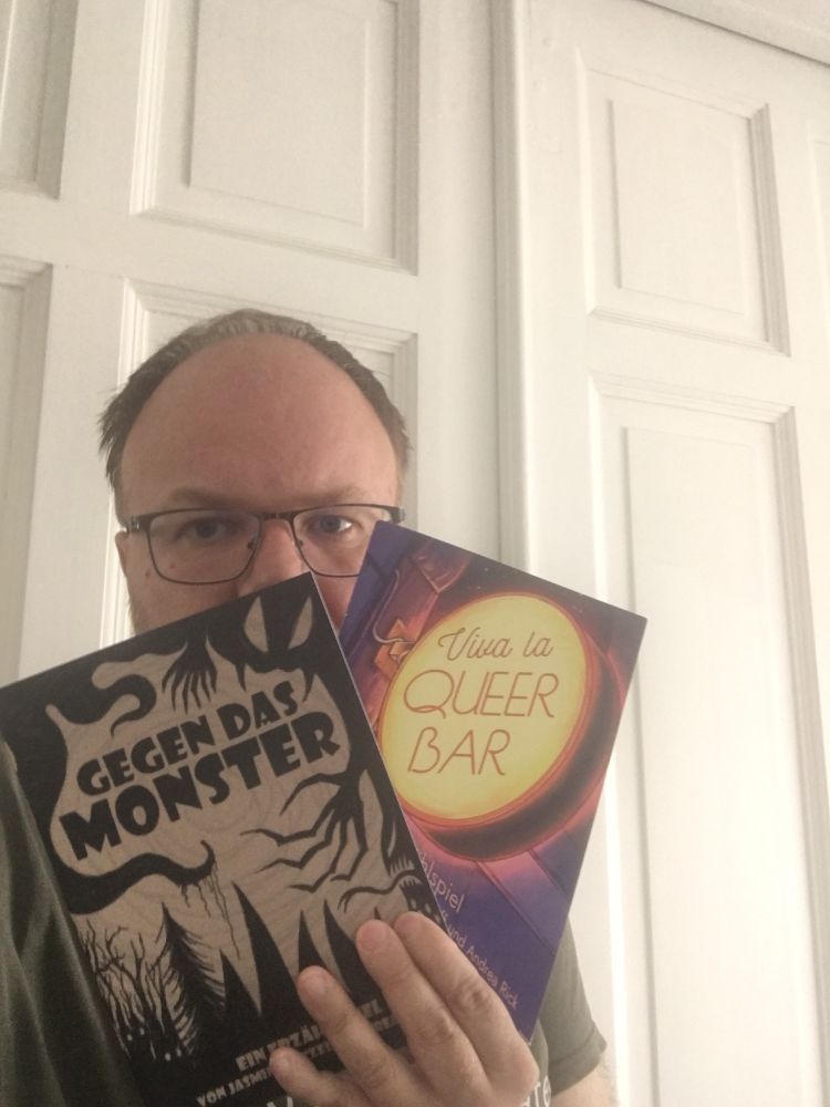 Me holding up two story games. Gegen das Monster is black and beige and has a creepy forest with tentacles and spidery fingers. Viva la Queer Bar has a neon sign saying Viva la Queer Bar.