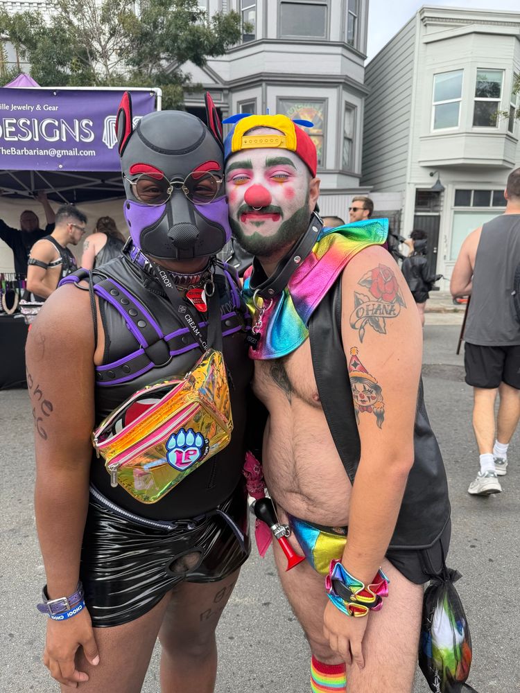 A nearly naked clown boy and a service dog share a tender moment on the street. On the left black, red, and purple hooded dog in a sleeveless body suit and short shorts, on the left the clown boy in a rainbow collar And matching jockstrap and leather vest, wearing eggshell white makeup with blush and red lipstick and a propeller hat 
