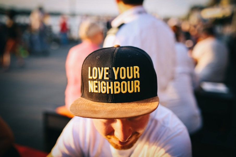 Young man in a white t-shirt, seated in a crowded public place on a sunny day, shows off his trucker cap. It reads: "Love your neighbor".