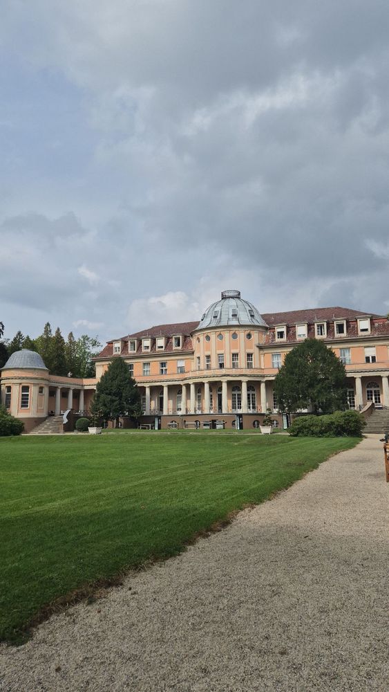 Außenansicht des Veranstaltungsorts: Ein historistisches Gebäude mit rosa Fassade, Säulengängen und einer Kuppel, flankiert von Rundbauten. Davor erstreckt sich ein großer grüner Rasen mit Kiesweg unter einem bewölkten Himmel. Es handelt sich um den Konzertsaal der BSP Business & Law School in Berlin, in dem die Tagung stattfindet.