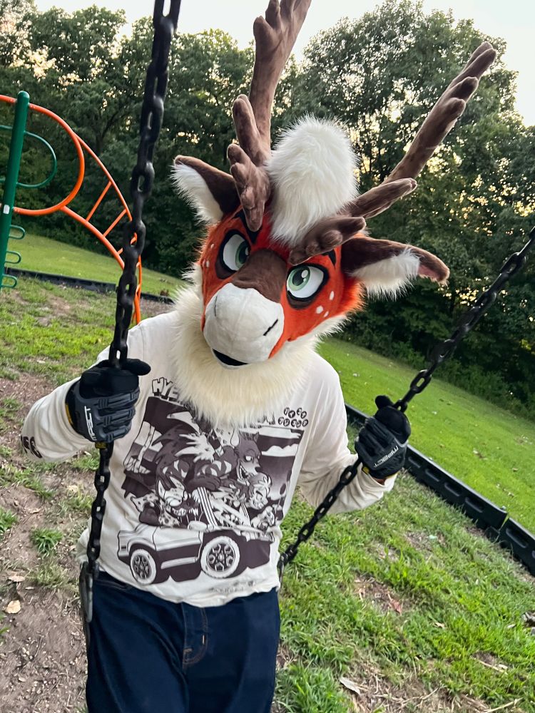 Caribou fursuiter in jeans and a graphic shirt on a swingset at a park