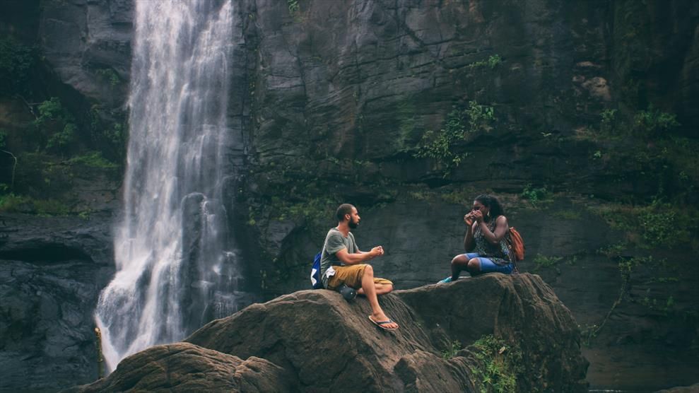 Two people sit together while smiling next to a waterfall.