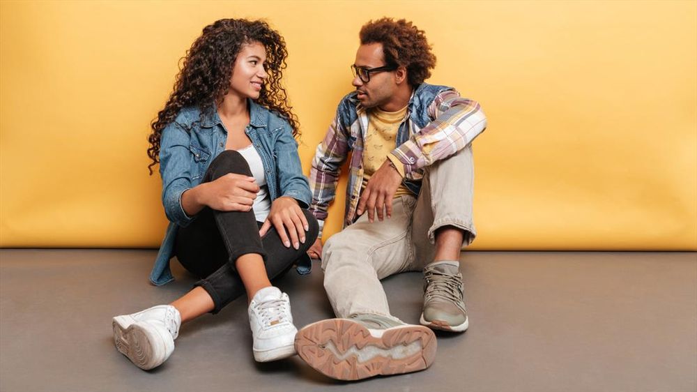 Two young people sitting on the floor against a yellow backdrop, dressed casually in denim and plaid shirts.