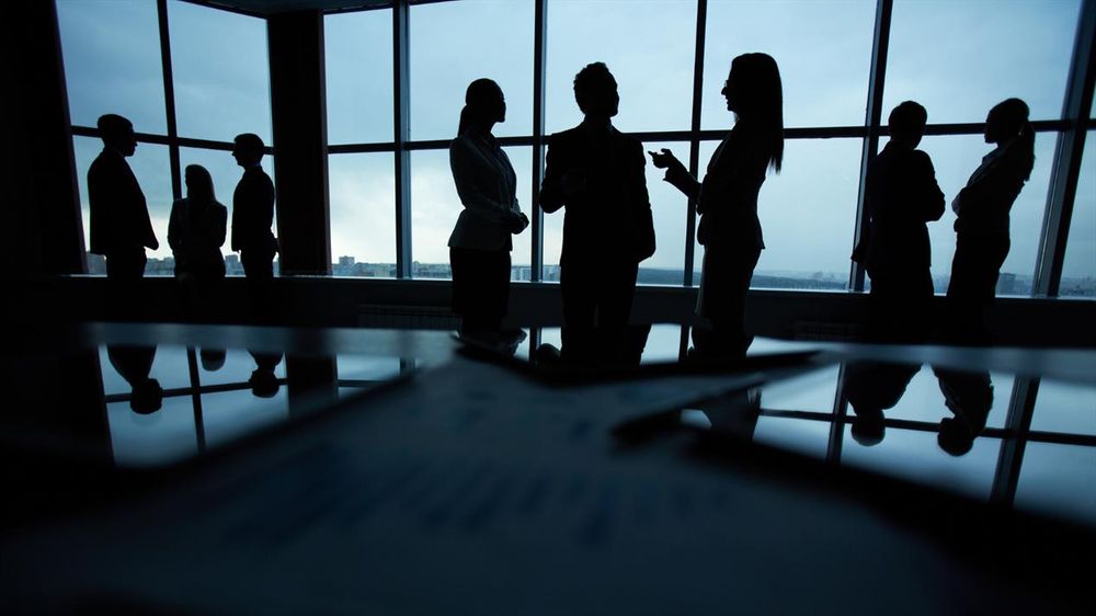 Business professionals in formal attire engaged in discussions near large office windows with city views at dusk.