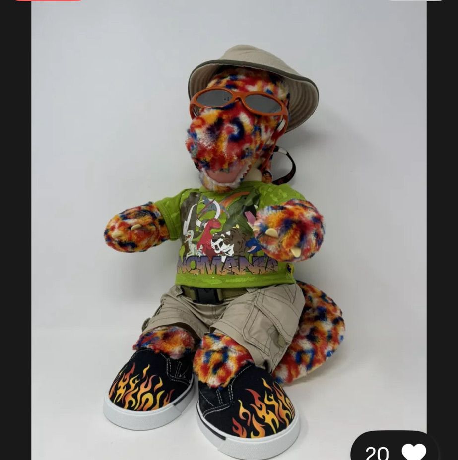 A fully dressed build a bear sitting on a white background. It's a rainbow confetti colored dinosaur dressed like a very cool 3rd grader: a bucket hat, sunglasses, a "Dinomania" T-shirt covered in prehistoric animals, cargo shorts, and Vans-type sneakers covered in flames. 