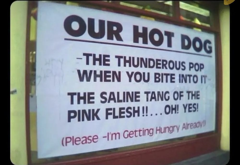 A white sign on a window that reads "OUR HOT DOG, THE THUNDEROUS POP WHEN YOU BITE INTO IT. THE SALINE TANG OF THE PINK FLESH!!...OH! YES! 

(Please, I'm getting hungry already!)"
