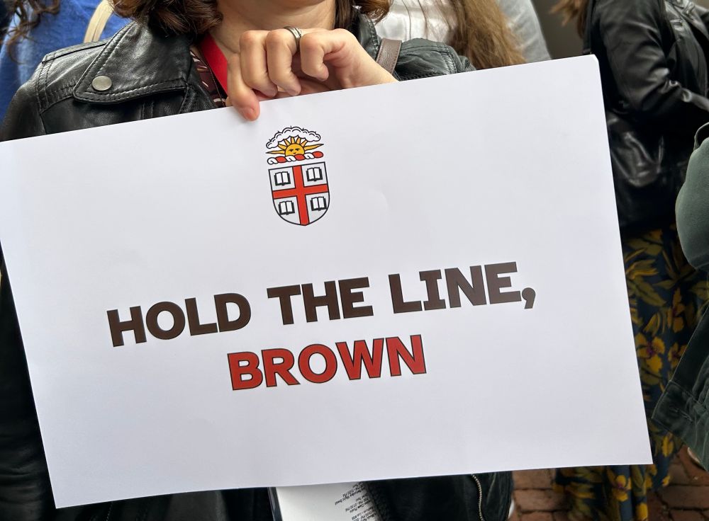 A hand holding a sign with Brown University’s crest and the text HOLD THE LINE, BROWN 