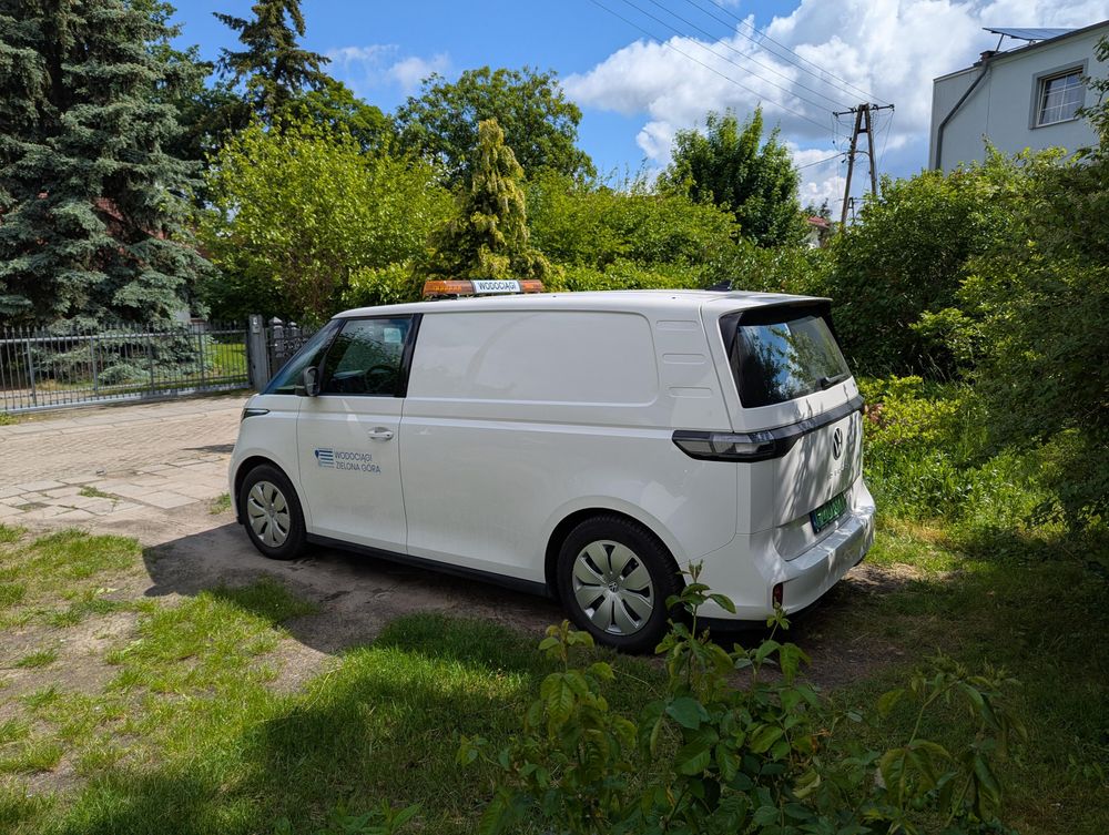 Biały elektryczny vanVW ID.Buzz widziany z boku. Na dachu kogut wodociągów. Tło - zielone krzaki.