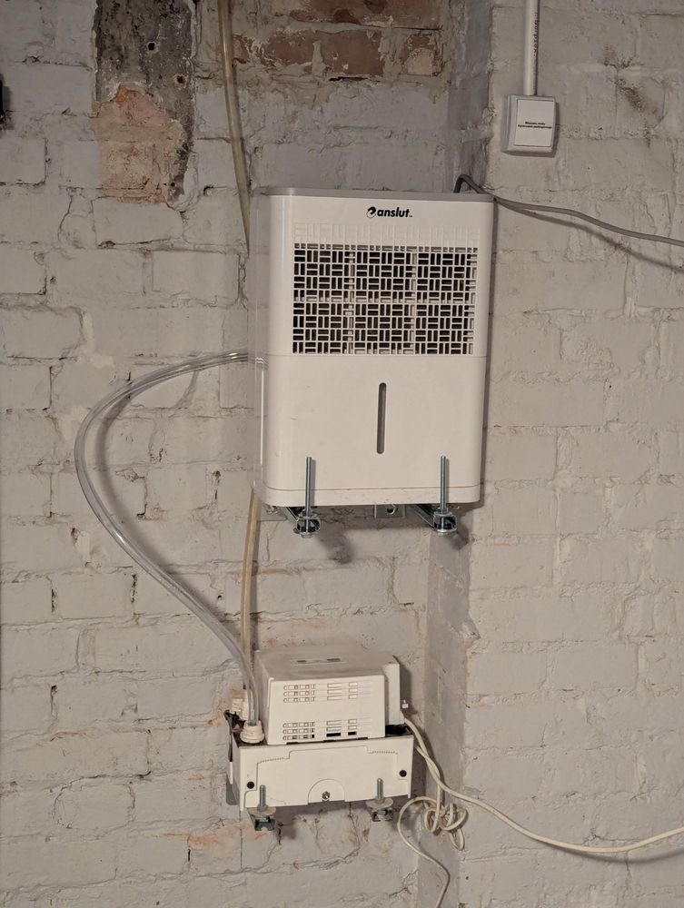 Dwa urządzenia elektryczne w białych obudowach zawieszone na ścianie. Wyżej osuszacz powietrza Anslut, od niego przezroczysty wężyk prowadzi do pompy zanurzeniowej która odprowadza wodę w górę do kanalizacji.