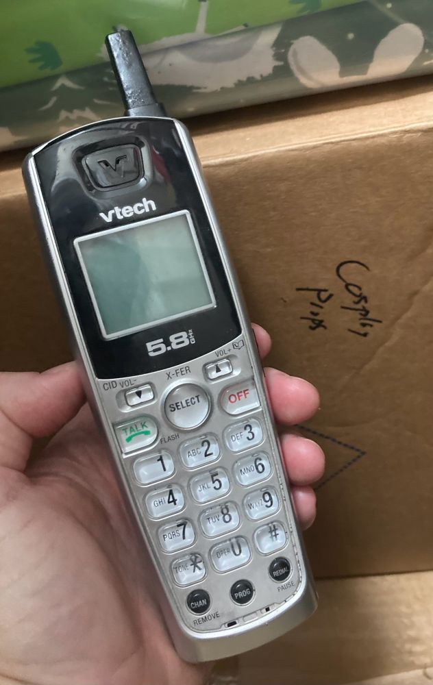 A cordless telephone being held in front of a box that says “cosplay props.”