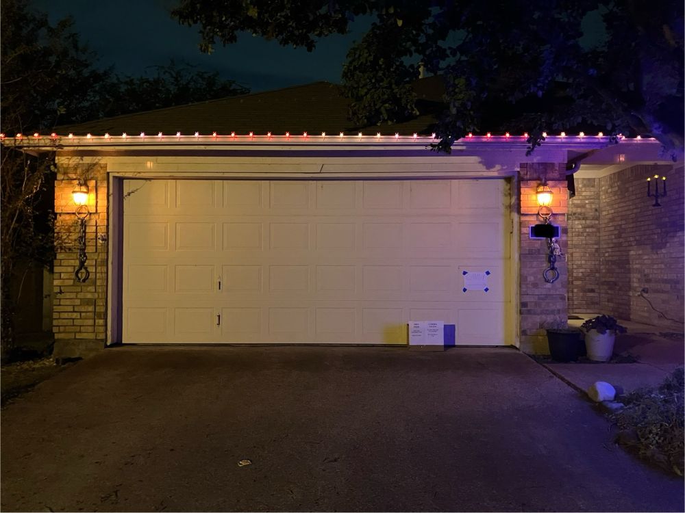 Garage door with flame lights on each side. Chains hang from the lights. Squirrel skeleton hangs from the chain on the left. A box with free food sits in the drive. 