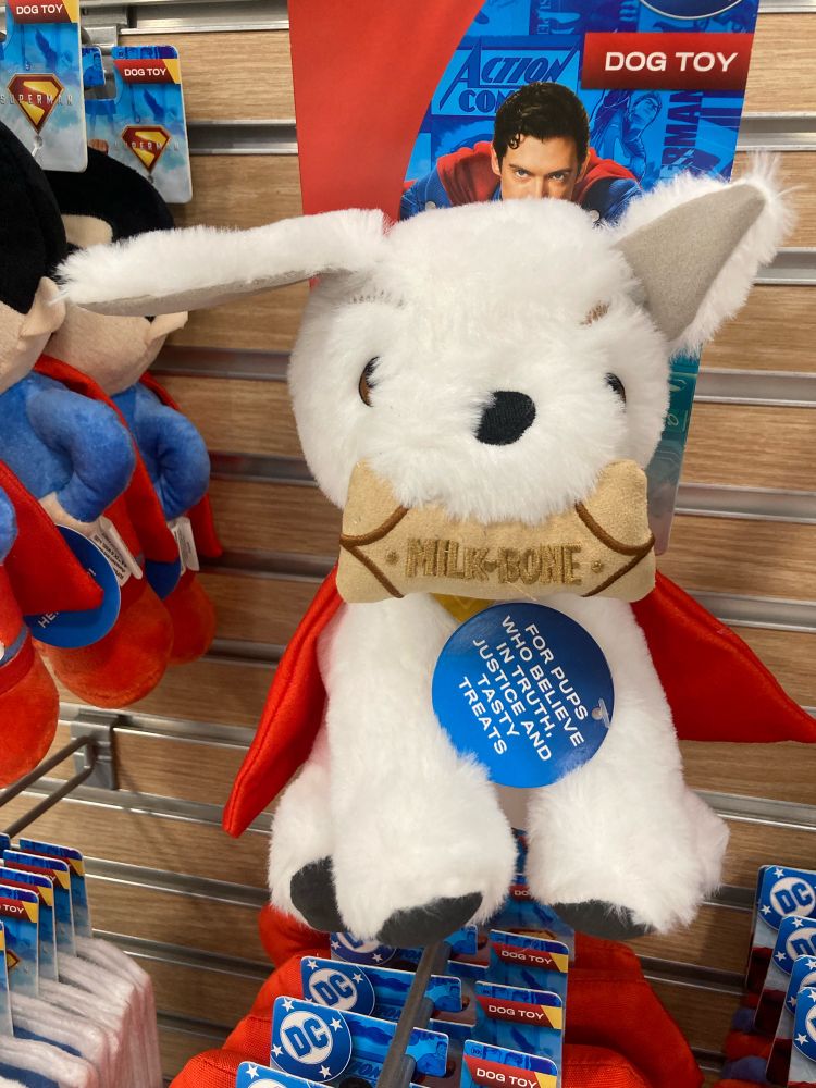 Stuffed Krypto toy. The collar tag reads: For pups who believe in truth, justice, and tasty treats. Krypto has a milk bone dog treat in his mouth.