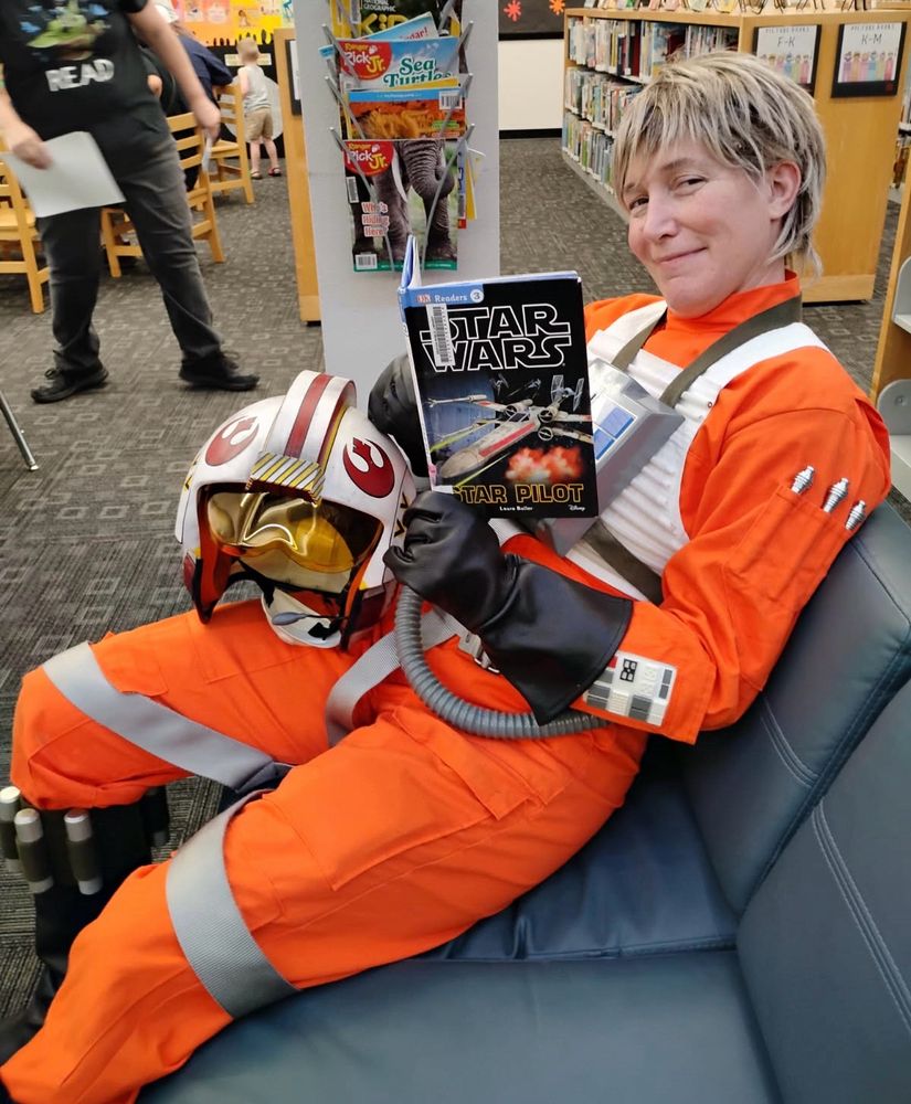 A Red Five Luke Skywalker cosplayer holding a book titled “Star Pilot” and grinning a smarmy grin at the camera. He is in his full flight suit with his helmet on his right thigh.