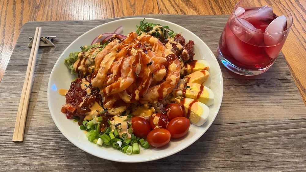A poke bowl made with romaine lettuce, brown rice, and topped with salmon "noodles." Arranged around the edges clockwise from the top are seaweed salad banchan, spicy squid banchan, hard-boiled egg slices, grape tomatoes, green onion, spicy octopus banchan, mashed avocado, and red onion. The top is drizzled with sriracha mayo and gochujang and sprinkled with furikake. To the left is a pair of disposable chopsticks propped up on a holder made from their wrapper, and on the right is a glass of mixed berry juice with ice.