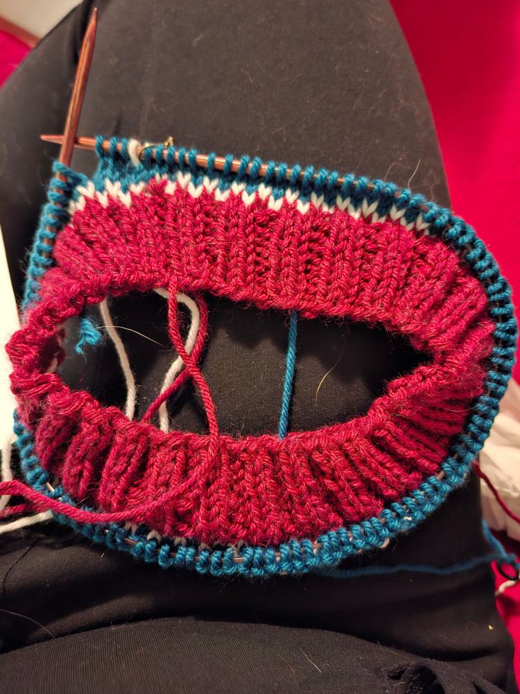 Knitting on the needles of cranberry red, white, and peacock blue.