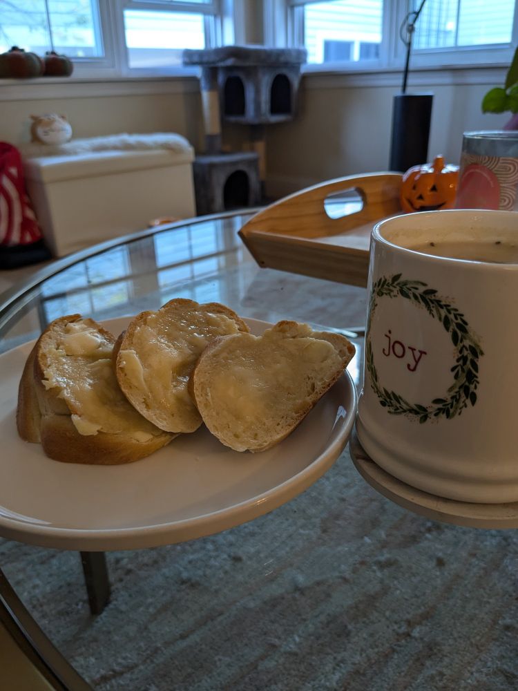 3 slices of bread slathered with honey butter on a white plate, next to a white mug that reads "joy" in red letters filled with tea. The tea and the mug are on a glass table and in the background is a cat tree and windows.
