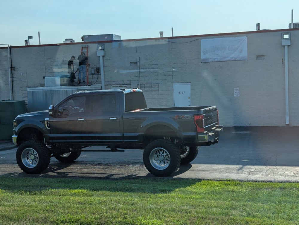 A big truck. Behind the big truck two men are trying to climb onto the roof of a building using only a short stepladder