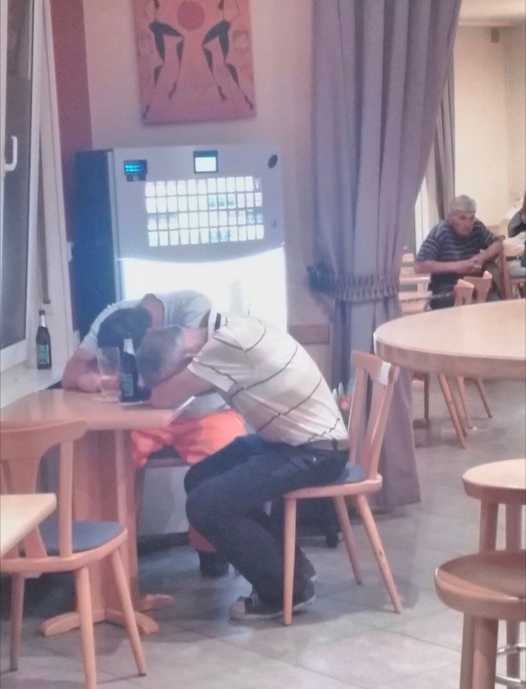 Two men seated at a table, slumped forward, possibly asleep, possibly dead, heads in arms. In the background, a man with a thousand yard stare drinks alone.